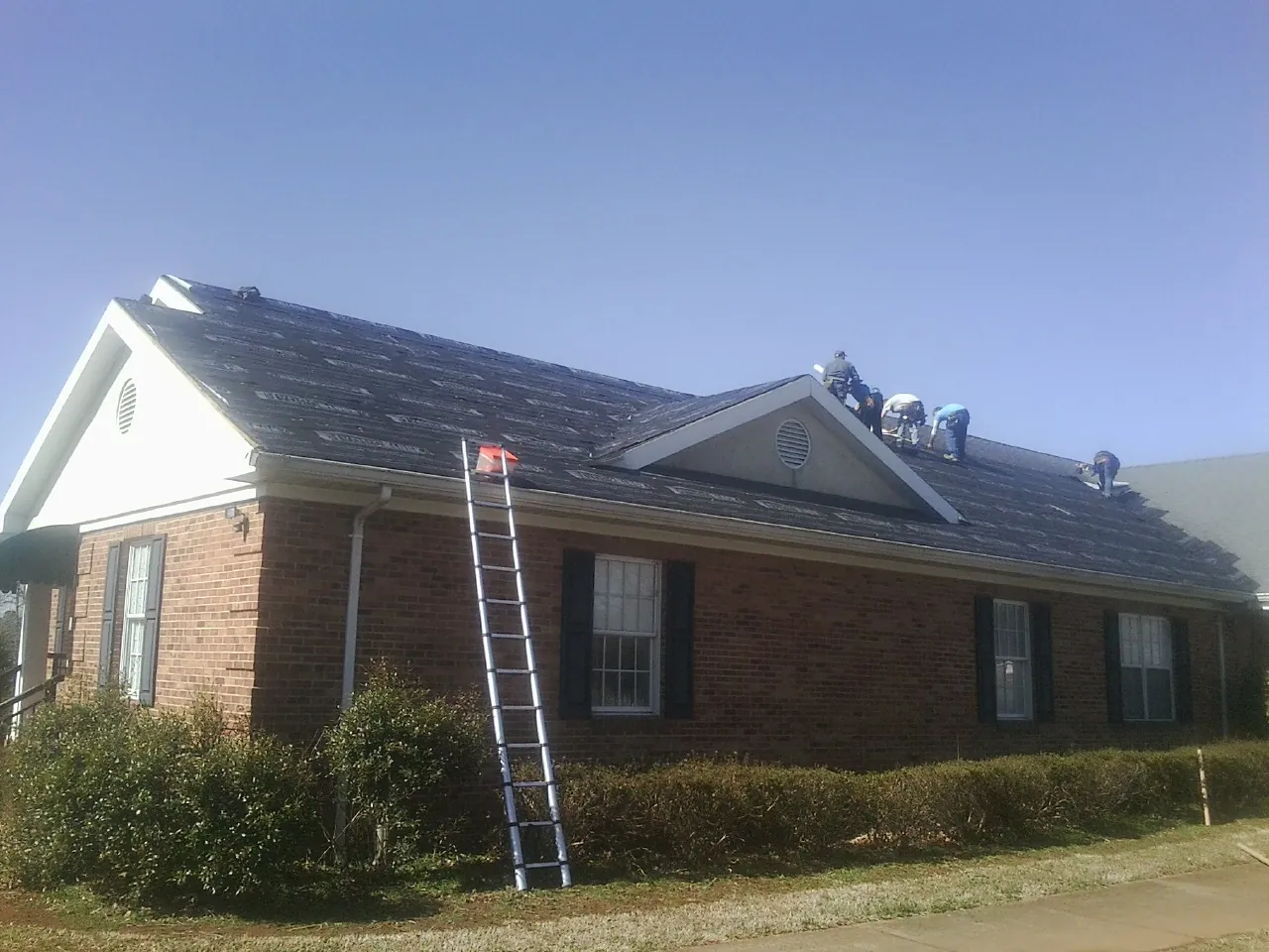 Crew tearing shingles off a brick ranch roof with a ladder set — Emergency Roof Repair work by our crew in Grifton