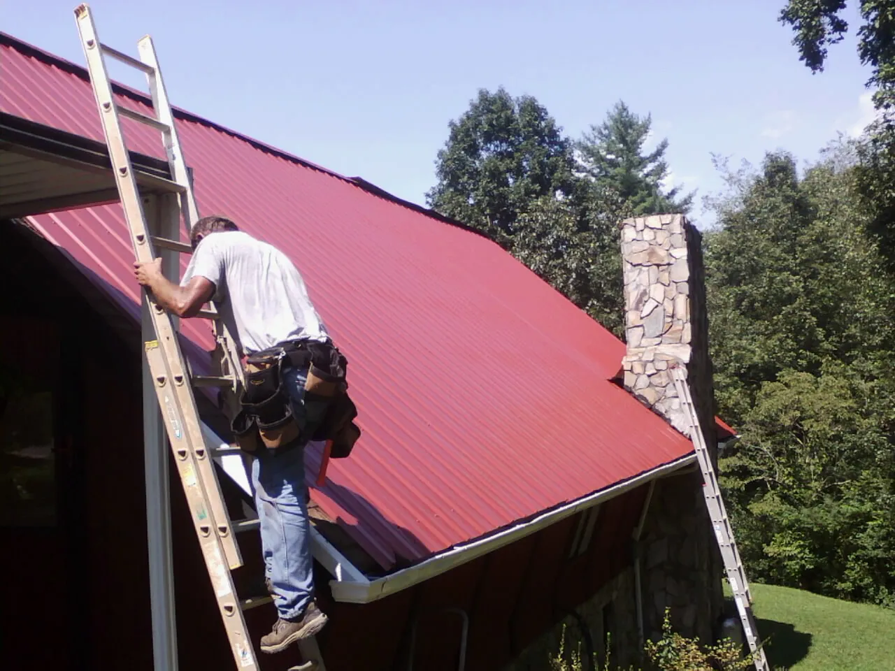 Roofer on a ladder beside a red metal roof and stone chimney — Soffit & Fascia Repair by Roofing Brothers in Grifton